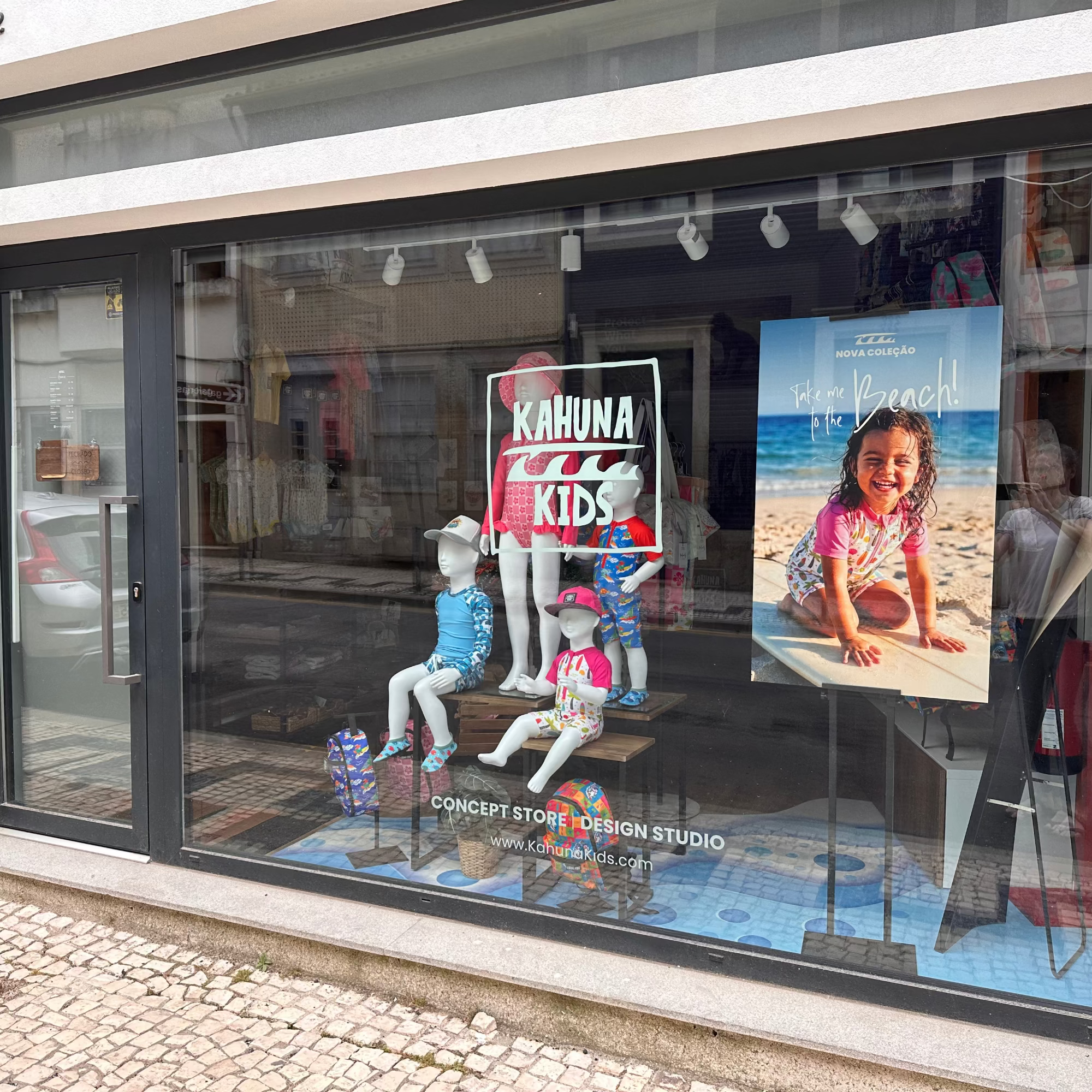 A storefront window display for Concept Store Vision Studio featuring mannequins in swimwear and a large poster of a child on a beach, alongside a 'Kahuna Kids' sign.