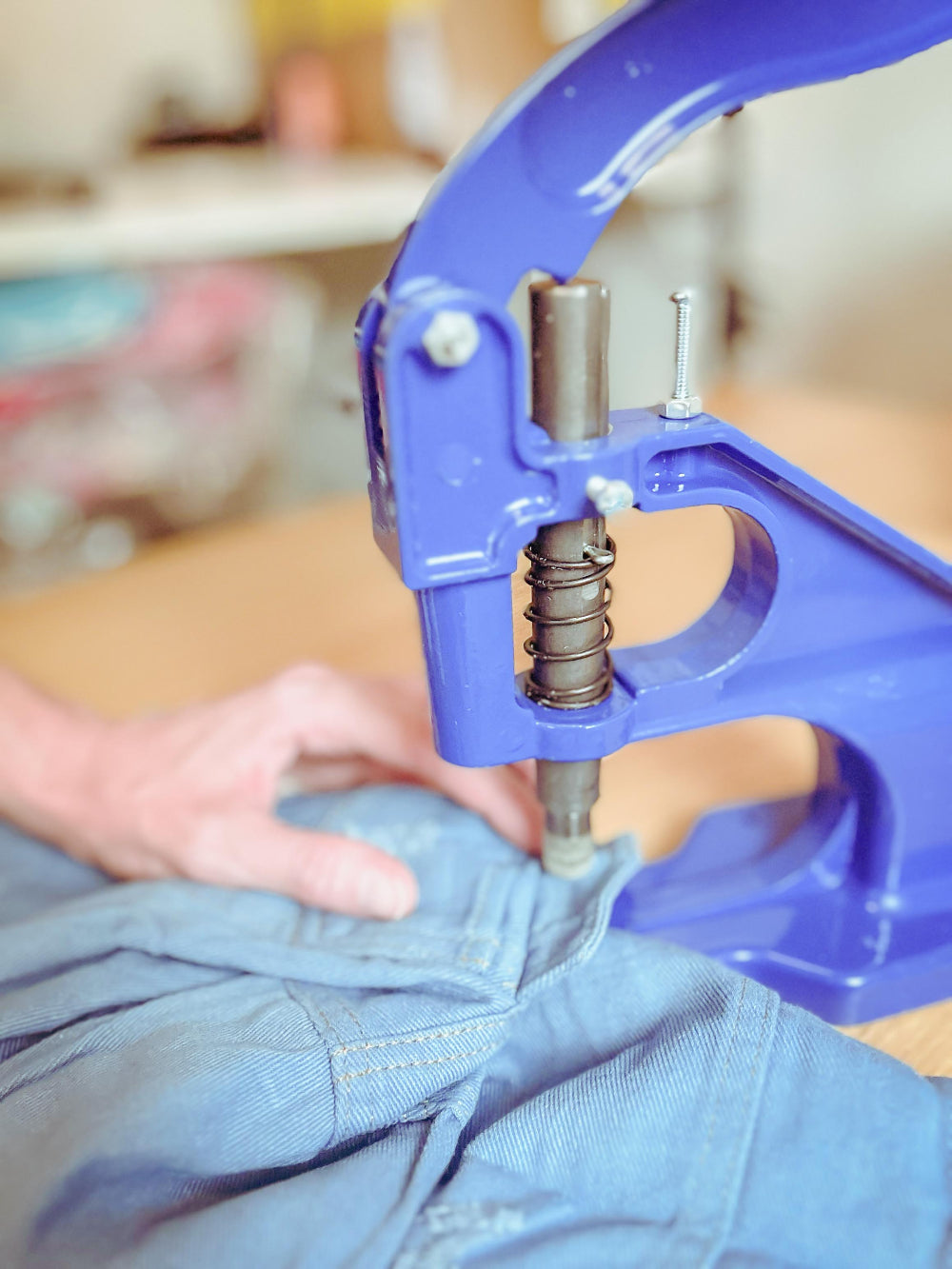 A close-up of a person's hand using a bright blue manual button-hole puncher on light blue denim jeans.