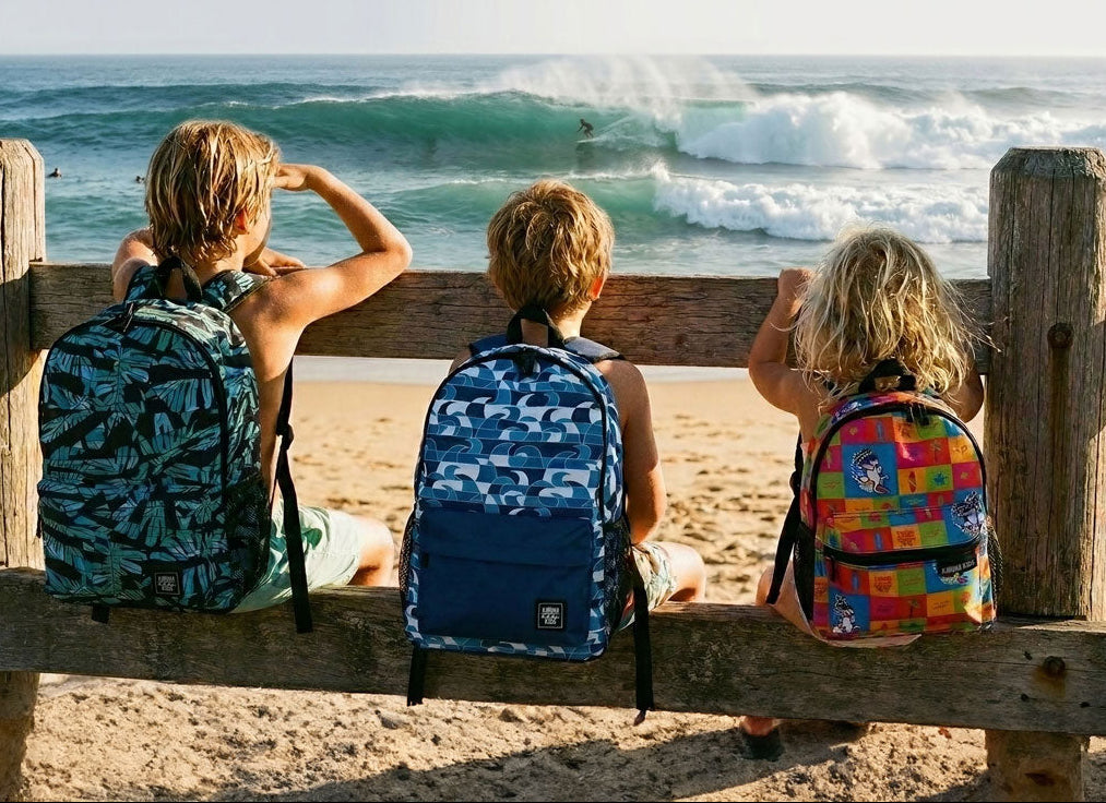 Image of Three children sitting on a wooden beach fence looking out at the ocean waves, each wearing a colorful patterned backpack. The scene is set on a sandy beach with the sea in the background.